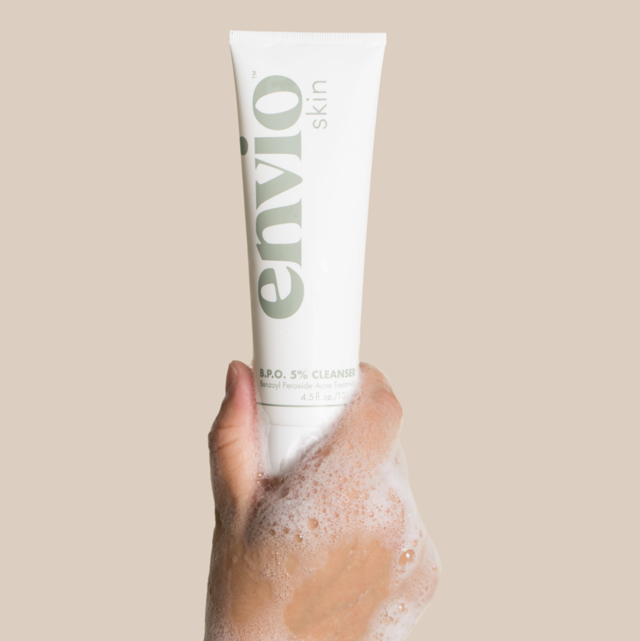 Envio Acne Cleanser Product Being Held Up By A Hand Covered In Soap Suds over a light brown background