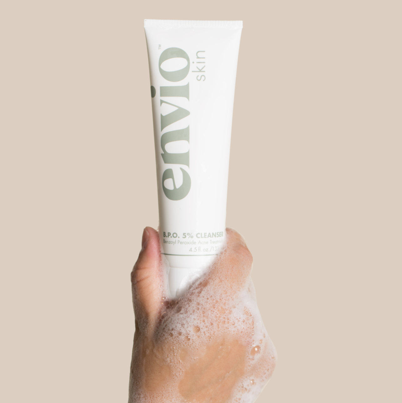 Envio Acne Cleanser Product Being Held Up By A Hand Covered In Soap Suds over a light brown background 